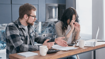 Stressed couple sitting at a kitchen table with financial documents, a calculator, and a laptop, illustrating the emotional and financial burden of managing debt or unexpected expenses.