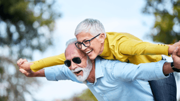 Joyful senior couple enjoying an outdoor moment, with the man playfully carrying the woman on his back, symbolizing happiness, security, and a carefree retirement.