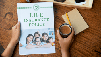 Hands holding a life insurance policy document featuring a smiling family on the cover, placed on a wooden table alongside a notebook, pencil, and cup of coffee, symbolizing financial planning and security.