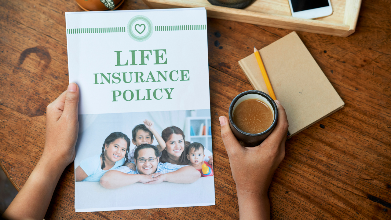 Hands holding a life insurance policy document featuring a smiling family on the cover, placed on a wooden table alongside a notebook, pencil, and cup of coffee, symbolizing financial planning and security.